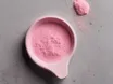 A pale pink scoop filled with pink powder on a grey concrete surface, with a small heap of the same powder beside it