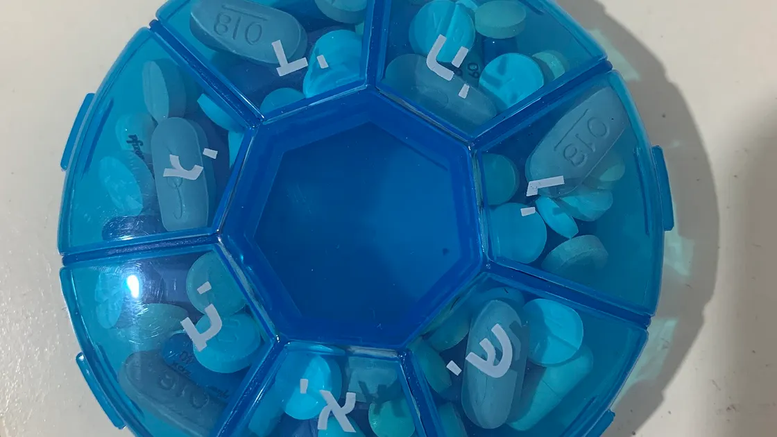 A blue seven-compartment weekly pill box, each compartment labelled with a Hebrew day letter and filled with assorted tablets